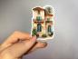 Preview: Ein handgehaltener Sticker, der eine mediterrane Villa mit einer warmen sandfarbenen Fassade, Palmen, einem Balkon und großen Fenstern zeigt. Die Villa ist im eleganten mediterranen Stil gestaltet, mit grünen Pflanzen im Vordergrund und einem einladenden,