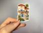 Preview: Ein handgehaltener Sticker, der eine mediterrane Villa mit einer orangefarbenen Fassade, einem Balkon mit schmiedeeisernem Geländer und großen, gewölbten Fenstern zeigt. Die Villa wird von einer Treppe und tropischen Pflanzen, darunter Palmen, umrahmt, wa