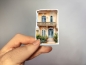 Preview: Aufkleber einer eleganten mediterranen Villa mit beiger Fassade, detailliertem Balkon und blauen Doppeltüren. Bild B.