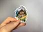 Preview: Sticker einer rustikalen Holzhütte in den Alpen, umgeben von grünen Bäumen, mit einem steinigen Pfad und schneebedeckten Berggipfeln im Hintergrund.