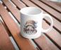 Preview: Weiße Tasse mit einem süßen Gnommotiv, abgebildet auf einer Holzbank im Freien. Der Gnom trägt eine gestrickte Mütze und ist umgeben von Bastelmaterialien wie Perlen und Nadeln. Ein charmantes Design für Handarbeits- und Bastelfans.