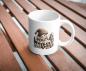 Preview: Weiße Tasse auf einem Holztisch mit einer Illustration eines kleinen, bärtigen Handwerkergnoms in einer gestrickten Mütze. Der Gnom sitzt an einem Tisch mit kleinen Skulpturen und Handwerksmaterialien, umgeben von weiteren Gnomenfiguren, die Liebe zum Det