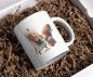 Preview: Weißer Keramikbecher mit einem Bild von einem Gnom in bunter Mütze, der auf einer Staffelei ein abstraktes, farbenfrohes Kunstwerk malt. Der Becher liegt auf braunem Füllmaterial in einer Geschenkbox, ideal als kreative Geschenkidee.