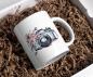 Preview: Weißer Keramikbecher in Geschenkverpackung mit einer Aquarellillustration einer Vintage-Kamera mit rosa Blüten, die sich um das Objektiv ranken. Der Becher liegt in einer naturfarbenen Papierpolsterung und verleiht dem Produkt eine rustikale und elegante