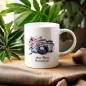 Preview: Weißer Keramikbecher mit einer Aquarellillustration einer Vintage-Kamera, umgeben von rosa Blüten, und dem Namen 'Susanne' darunter. Der Becher ist festlich arrangiert mit einem Zweig Tannenzweigen und goldenen Weihnachtskugeln im Hintergrund, was eine el