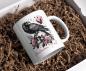Preview: Weiße Tasse mit Halloween-Design: Ein schwarzer Rabe sitzt auf einem Totenkopf, umgeben von rosafarbenen Rosen und Blättern. Im Hintergrund fliegt ein weiterer Rabe vor einem rosa Mond. Der Schriftzug 'Halloween y'all' in elegantem Schriftstil rundet das