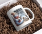 Preview: Weihnachtstasse mit Schneemann, der am Fenster steht und eine Tasse Kakao hält, umgeben von Tannenzapfen und Plätzchen. Die Tasse vermittelt winterliche Gemütlichkeit und ist perfekt für die Festtagssaison.