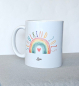 Preview: Weiße Keramiktasse mit pastellfarbenem Regenbogen, Herzchen und dem Schriftzug „Schulkind 2025“ in Hellblau, fotografiert auf einem Holztisch.