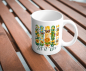 Preview: Weiße Keramiktasse mit farbenfrohem Naturmotiv aus Sonnenblumen, Bienen, Herzen und Schmetterlingen sowie dem Schriftzug „Let it Bee“, fotografiert auf einem Holztisch.