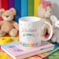Preview: Personalisierte Tasse „Schulkind 2026“ mit buntem Schulanfang-Motiv, farbigen Stiften, Regenbogen und Name „Bruno“ in Blau, dekoriert mit Büchern und Kuscheltieren im Hintergrund.