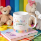 Preview: Personalisierte Tasse „Schulkind 2026“ mit buntem Schulanfang-Motiv, farbigen Stiften, Regenbogen und Name „Leona“ in Rosa, dekoriert mit Büchern und Kuscheltieren im Hintergrund.