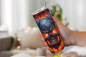 Preview: Hand hält einen Thermobecher mit Totenkopf-Design, das feurige und leuchtende Blau- und Rottöne zeigt. Der Becher hat einen silbernen Deckel mit Strohhalm. Im Hintergrund ist eine unscharfe, gemütliche Wohnzimmerszene mit warmem Bokeh-Lichteffekt.
