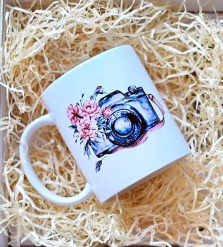 Handgefertigte weiße Tasse mit einer Kamera-Illustration und floralen Elementen. Perfekt als Geschenk für Fotografen, präsentiert in einer dekorativen Holzwolle-Verpackung.