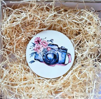 Runder Untersetzer mit einer Illustration einer Kamera und Blumen im Aquarellstil, dekorativ in Holzwolle verpackt.