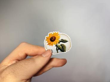 Hand, die einen Aufkleber in Form einer Sonnenblume hält, gestaltet von DreamArt VM.