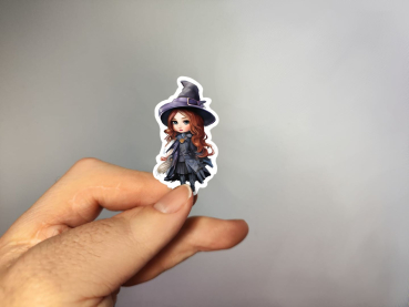 Hand, die einen Aufkleber in Form einer kleinen Hexe mit langen roten Haaren, schwarzem Hut und schwarzem Kleid hält, gestaltet von DreamArt VM.