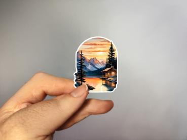 Aufkleber mit einer malerischen Hütte am Ufer eines ruhigen Sees bei Sonnenuntergang. Im Hintergrund erheben sich majestätische Berge, während der Himmel in warmen Orange- und Gelbtönen leuchtet. Hohe Tannenbäume umrahmen die Szene und das Wasser spiegelt