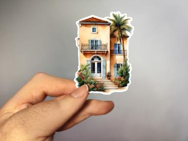 Ein handgehaltener Sticker, der eine elegante mediterrane Villa mit hellgelber Fassade, blauen Fensterläden und einem Balkon zeigt. Vor der Villa befindet sich eine Treppe, die von tropischen Pflanzen und Palmen umgeben ist, was dem Gebäude einen exotisch