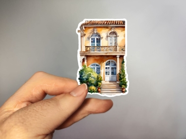 Aufkleber einer mediterranen Villa mit zwei Etagen, cremefarbener Fassade und Balkontüren, flankiert von grünen Büschen. Bild C.