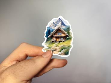 Sticker einer rustikalen Berghütte inmitten einer malerischen Alpenlandschaft mit einem schneebedeckten Berg im Hintergrund, umgeben von grünen Wiesen und Nadelbäumen.
