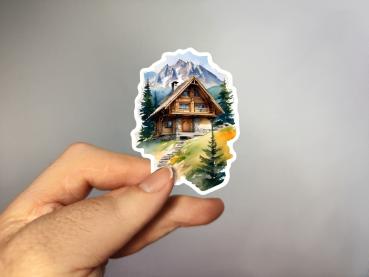 Sticker einer traditionellen Berghütte mit Holzfassade und Steinfundament, umgeben von Tannenbäumen und Bergen im Hintergrund.