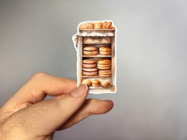 Hand hält einen Aufkleber, der eine Vitrine mit verschiedenen frischen Brötchen und Gebäck zeigt, Bild A. Die Illustration ist im handgezeichneten Vintage-Stil gehalten und zeigt Gebäck auf mehreren Regalen, ordentlich gestapelt.