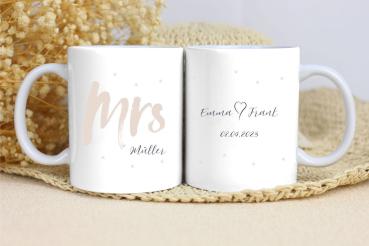 Zwei weiße Keramikbecher nebeneinander auf einer beigen Unterlage mit Trockenblumen im Hintergrund. Der linke Becher zeigt den Text 'Mrs Müller' in beiger Farbe mit kleinen Punkten als Dekoration. Der rechte Becher trägt den Schriftzug 'Emma ♥ Frank' mit