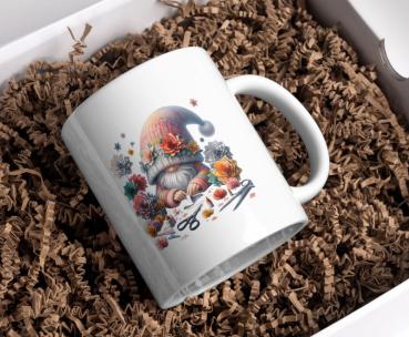 Weißer Kaffeebecher mit einem niedlichen Gnom-Design, umgeben von bunten Blumen und Bastelutensilien, präsentiert in einer Geschenkbox mit Papierfüllmaterial. Perfekt als Geschenk für kreative Seelen und Gartenliebhaber.