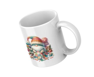 Ein schräg stehender weißer Keramikbecher hat ein farbenfrohes Design. Der Becher zeigt ein Bild von einem fröhlichen Zwerg, der in einem bunten Deckenstück eingewickelt ist. Um ihn herum sind Wollknäuel, Nähutensilien und andere kreative Materialien, die