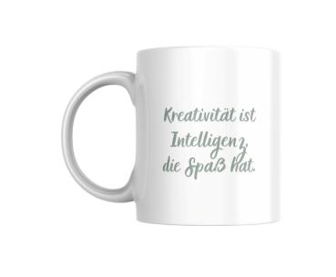 Ein weißer Keramikbecher steht auf einem einfarbigen Hintergrund. Der Becher ist mit einer inspirierenden Aufschrift verziert, die lautet: 'Kreativität ist Intelligenz, die Spaß hat.' Die Schrift ist in einem sanften Grünton gehalten und verleiht dem Bech