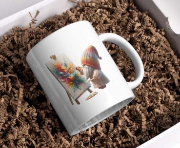 Weißer Keramikbecher mit einem Bild von einem Gnom in bunter Mütze, der auf einer Staffelei ein abstraktes, farbenfrohes Kunstwerk malt. Der Becher liegt auf braunem Füllmaterial in einer Geschenkbox, ideal als kreative Geschenkidee.