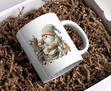 Weißer Keramikbecher mit einem Motiv eines kleinen Gnoms in Strickmütze und Pullover, der in einer Werkstatt an der Herstellung von handgemachter Seife arbeitet. Der Becher liegt in einer Geschenkbox auf braunem Füllmaterial und ist ideal für Liebhaber vo