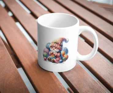 Weißer Keramikbecher mit einem Motiv eines niedlichen Gnoms in einer bunten Strickmütze, der mit farbenfrohen Garnknäueln strickt. Der Becher steht auf einem Holztisch und ist perfekt für Handarbeitsliebhaber und kreative Menschen.