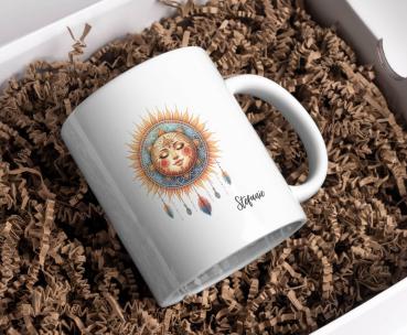 Weißer Keramikbecher mit einem Boho-Sonnenmotiv, das ein lächelndes Gesicht umgeben von Sonnenstrahlen und hängenden Federn zeigt, und dem Namen ‚Stefanie‘ unten rechts aufgedruckt. Der Becher liegt auf braunem Füllmaterial in einer Geschenkbox und ist ei