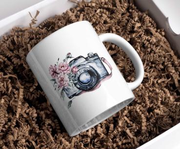 Weißer Keramikbecher in Geschenkverpackung mit einer Aquarellillustration einer Vintage-Kamera mit rosa Blüten, die sich um das Objektiv ranken. Der Becher liegt in einer naturfarbenen Papierpolsterung und verleiht dem Produkt eine rustikale und elegante
