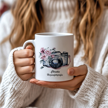 Preview: Personalisierte Fotografin-Tasse mit Aquarellkamera und rosa Blumenmotiv, ideales Geschenk für Fotografen, aufgenommen in einer gemütlichen Handszene.