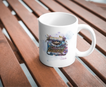 Weiße Keramiktasse mit kunstvoller Aquarell-Illustration einer Vintage-Schreibmaschine, verziert mit floralen Elementen und einem personalisierten Namen „Claudia“. Die Tasse steht auf einem Holztisch in natürlichem Licht und vermittelt eine gemütliche, in