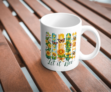 Weiße Keramiktasse mit farbenfrohem Naturmotiv aus Sonnenblumen, Bienen, Herzen und Schmetterlingen sowie dem Schriftzug „Let it Bee“, fotografiert auf einem Holztisch.