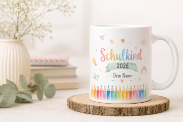 Personalisierte Tasse „Schulkind 2026“ mit buntem Schulanfang-Motiv, farbigen Stiften, Regenbogen und Platzhalter „Dein Name“, dekoriert auf einer Holzscheibe mit Büchern, Vase und Eukalyptus im Hintergrund.