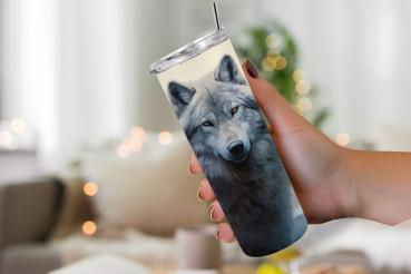 Hand hält einen Thermobecher mit einem detaillierten Wolfsportrait, das das Gesicht eines grauen Wolfs mit durchdringendem Blick zeigt. Der Becher hat einen silbernen Deckel mit Edelstahlstrohhalm. Im Hintergrund ist eine unscharfe, gemütliche Wohnzimmers