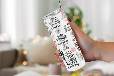 Hand hält einen weißen Thermobecher mit motivierenden Zitaten wie 'The future is mine', 'Good things are coming' und 'Keep going, keep growing'. Der Becher ist mit floralen Illustrationen und einem Regenbogen verziert. Im Hintergrund ist ein gemütlicher R