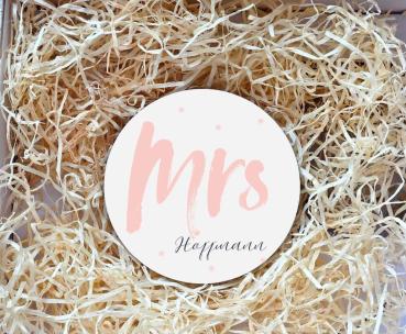 Runder Untersetzer mit dem Schriftzug „Mrs“ in rosafarbener Schrift und dem Nachnamen „Hoffmann“ darunter in eleganter Schreibschrift. Der Untersetzer liegt in einer Geschenkbox, umgeben von natürlichem Holzwolle-Füllmaterial. Perfekt als personalisiertes