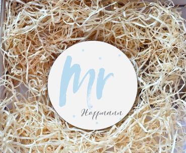 Runder Untersetzer mit dem Schriftzug „Mr“ in hellblauer Schrift und dem Nachnamen „Hoffmann“ darunter in eleganter Schreibschrift. Der Untersetzer liegt in einer Geschenkbox, umgeben von natürlichem Holzwolle-Füllmaterial. Perfekt als personalisiertes Ge