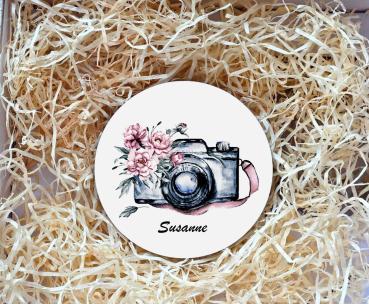 Runder Untersetzer mit einer Illustration einer Kamera und rosafarbenen Blumen sowie dem Namen „Susanne“ in eleganter Schrift darunter. Der Untersetzer liegt in einer Geschenkbox, umgeben von natürlichem Holzwolle-Füllmaterial. Perfekt als personalisierte