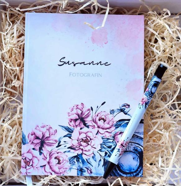 Personalisierter Notizblock mit Namen 'Susanne, Fotografin' und passendem Stift, beide mit floralen Motiven dekoriert, stilvoll in Holzwolle verpackt.