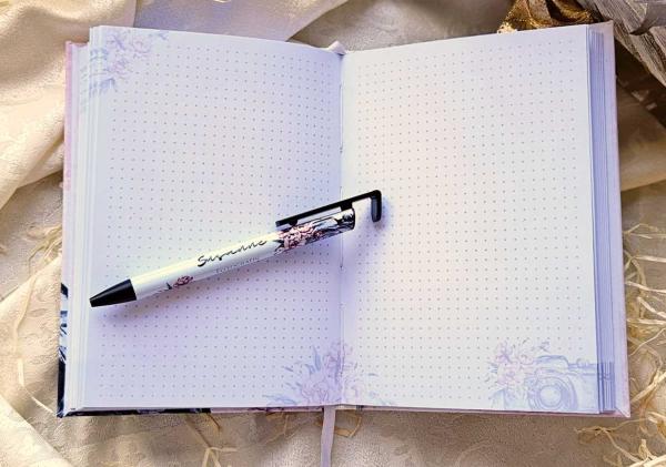 Offenes Notizbuch mit personalisiertem Kugelschreiber 'Susanne', gestaltet mit floralen und fotografischen Motiven, auf hellem Stoffhintergrund.