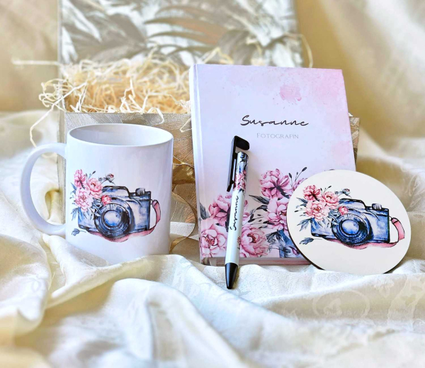 Fotografen-Geschenkset mit einer Tasse, einem Notizbuch, einem Stift und einem runden Untersetzer, alle mit floralen und Kameramotiven personalisiert und auf einem hellen Stoffhintergrund präsentiert.