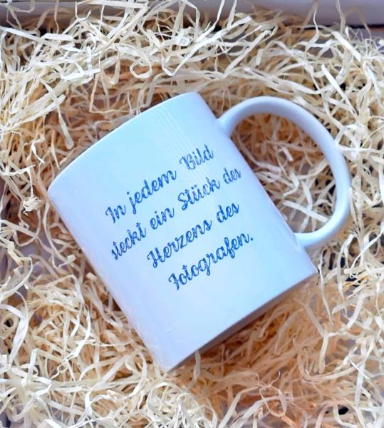 Handgefertigte weiße Tasse mit dem Schriftzug 'In jedem Bild steckt ein Stück des Herzens des Fotografen'. Präsentiert in einer dekorativen Holzwolle-Verpackung.