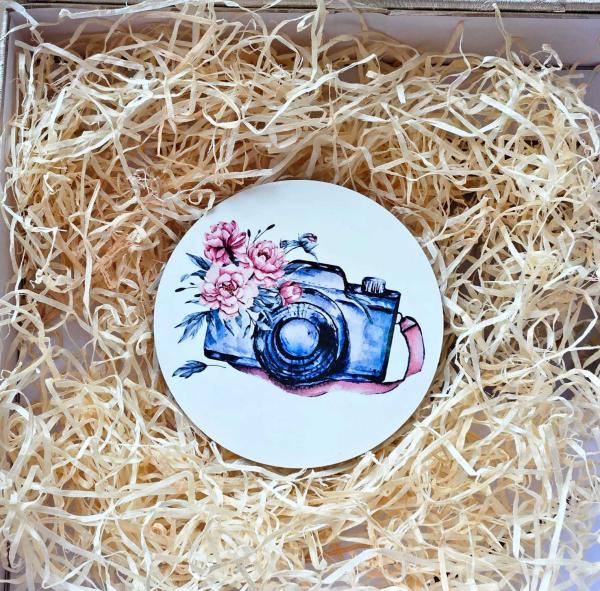 Runder Untersetzer mit einer Illustration einer Kamera und Blumen im Aquarellstil, dekorativ in Holzwolle verpackt.