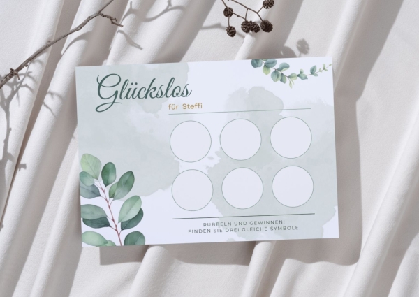 Personalisierte Glückslos-Karte mit dem Namen „Steffi“ im Eukalyptus-Design mit grünen Blättern und zartem Aquarellhintergrund. Die Karte liegt auf hellem Stoff neben einem Zweig und zeigt sechs leere Rubbelfelder.